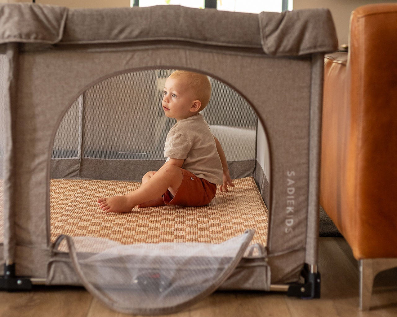 Child sitting inside a portable playpen with 'Sadek' branding in a home setting.